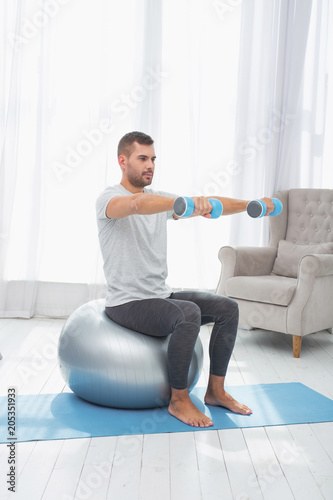 Sports equipment. Serious handsome man sitting on the med ball while exercising with dumbbells photo