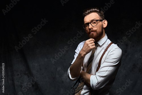 Young stylish hipster with cool hairstyle and beard dressed in white shirt and suspenders is thinking of a new creative idea looking at viewer