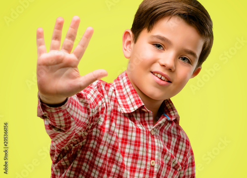 Handsome toddler child with green eyes raising finger, is the number five over yellow background