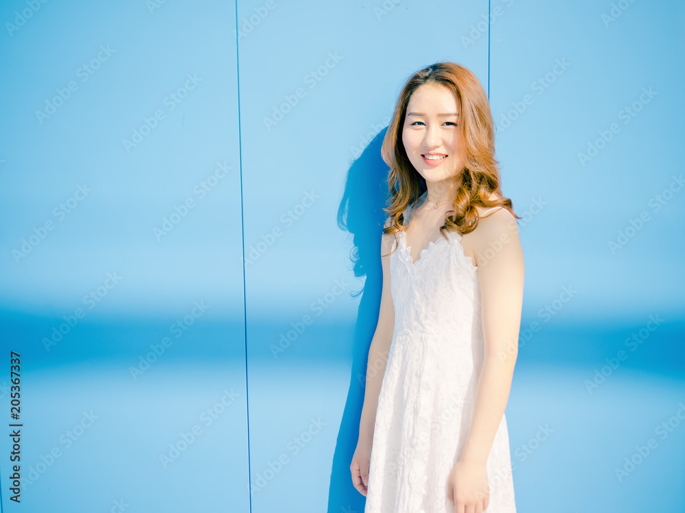 Beautiful Chinese brunette woman in white wedding dress posing and smiling at camera with blue wall background. Sunny portrait of glamour young stylish lady. Emotions, beauty and lifestyle concept.