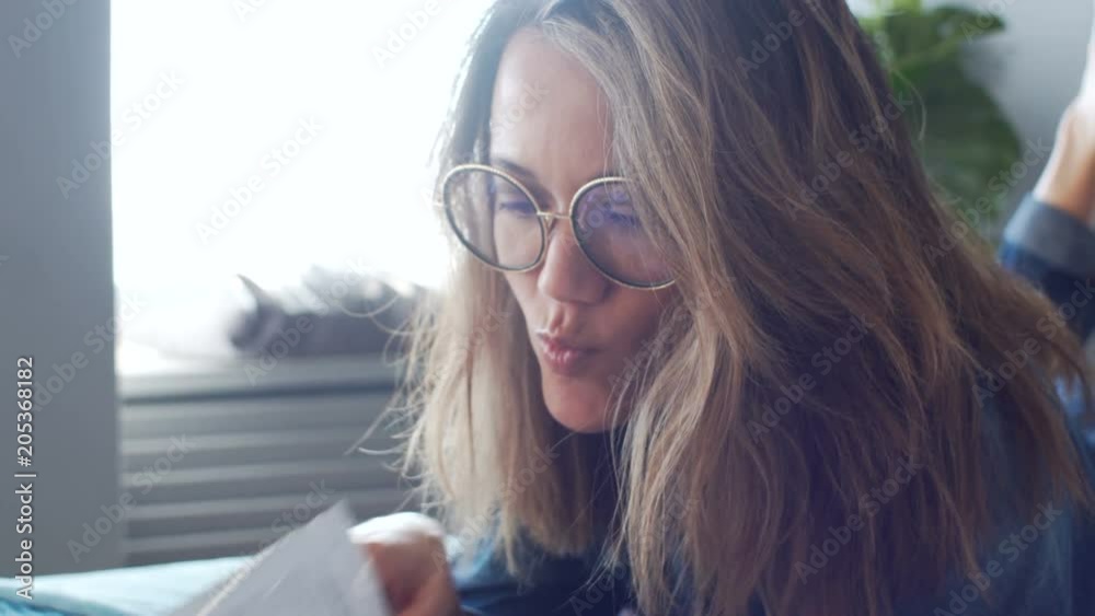 Close-up of attractive caucasian woman in twenties in round glasses reading and looking through glossy magazine in cozy bed during the morning in spacious stylish sunny bedroom with decor and textile Close-up of attractive caucasian woman in twenties in round glasses reading and looking through glossy magazine in cozy bed during the morning in spacious stylish sunny bedroom with decor and textile