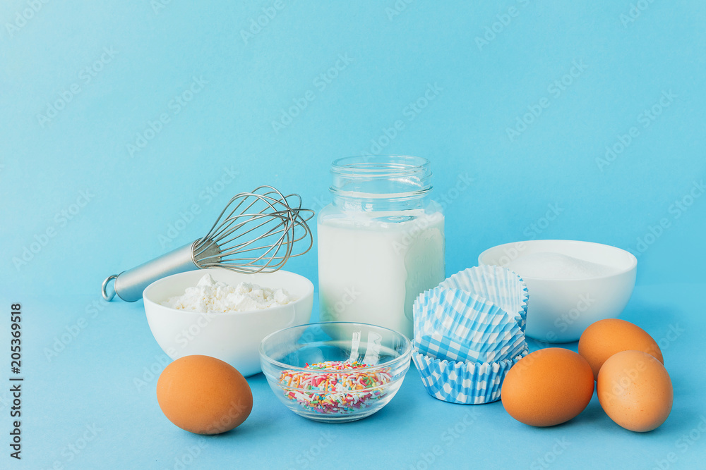 Eggs of chicken, flour in a bowl, sugar, milk in a glass jar whisk ingredient for cooking a bake on a blue background with copy space Eggs of chicken, flour in a bowl, sugar, milk in a glass jar whisk ingredient for cooking a bake on a blue background with copy space