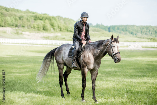Man in leather jacket with protective helmet riding a horse on the green meadow