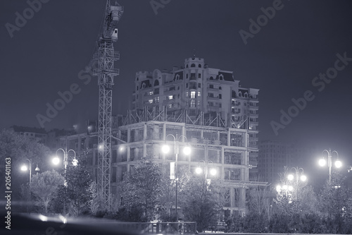 In the evening building is going to be built. Silhouette Building crane and building under construction against evening cloudy sky . Tall yellow cranes with illumination and buildings under © adilcelebiyev