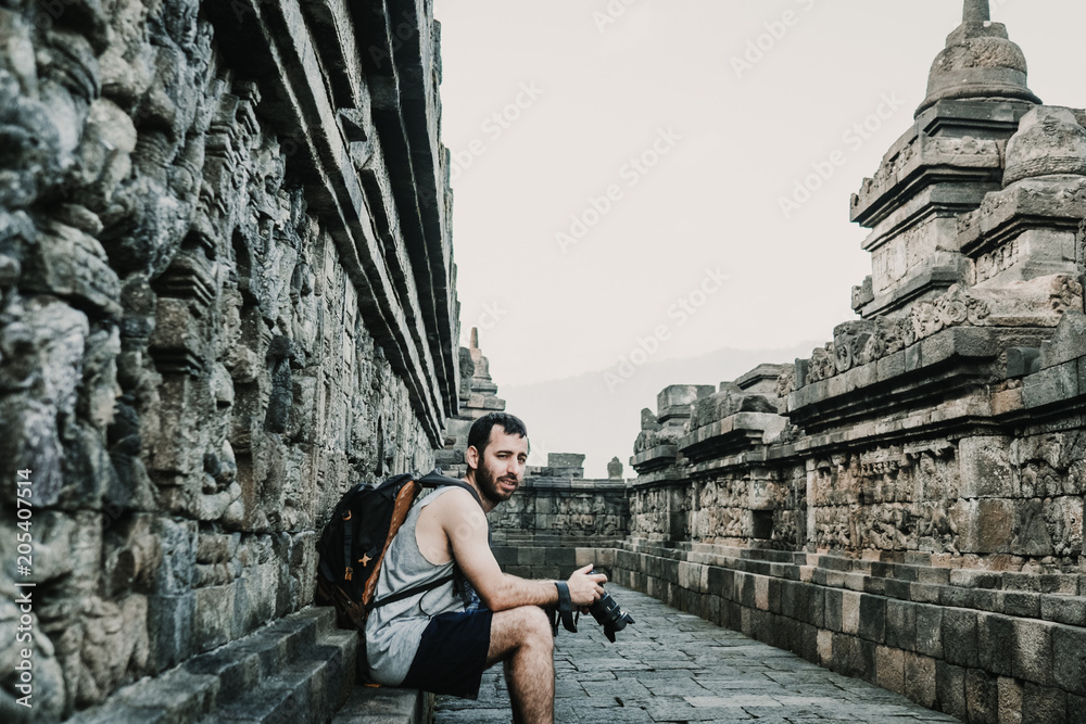 Handsome young tourist feeling the peace and taking some pictures of the great Borobudur temple, historical famous place in the java island Indonesia. Lifestyle and travel photography.