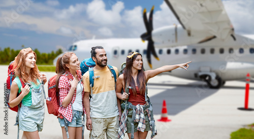 air travel, tourism and adventure concept - group of smiling friends with backpacks pointing finger to something over plane on airfield background