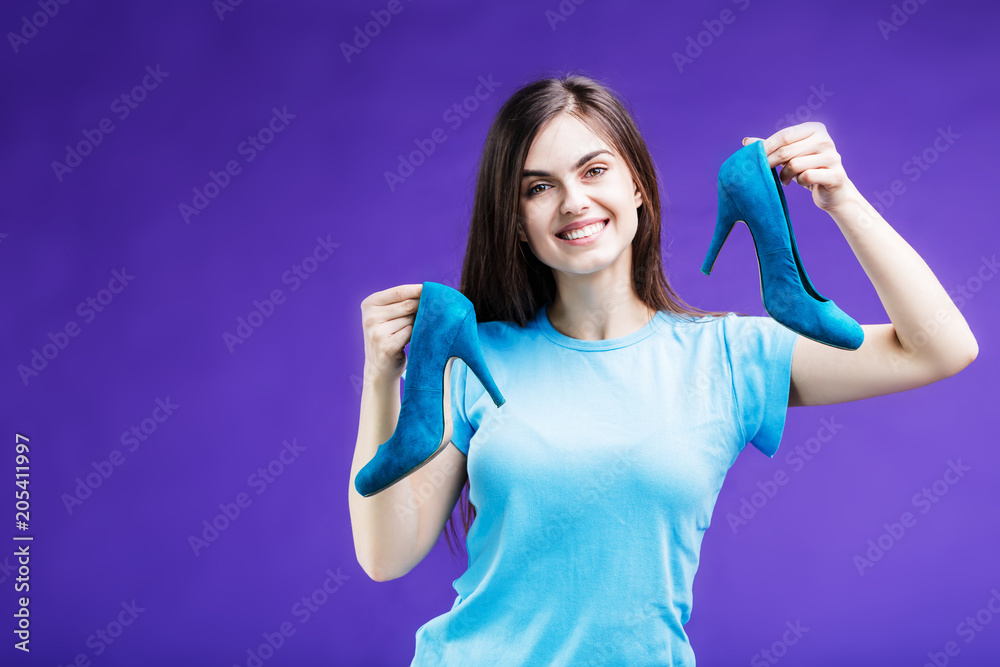 Pretty happy smiling brunette girl dressed in blue t-shirt holding teal heels shoes before blue background Pretty happy smiling brunette girl dressed in blue t-shirt holding teal heels shoes before blue background