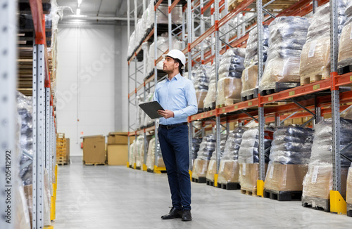 logistic business, shipment and people concept - businessman in helmet with clipboard checking goods at warehouse