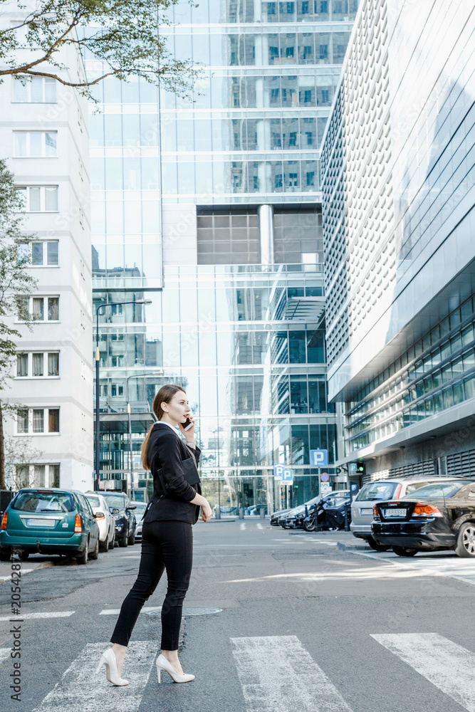 A young woman in a business suit crosses the street. Business woman talking through a mobile phone. A girl with a notepad and a phone near the office. A young woman in a business suit crosses the street. Business woman talking through a mobile phone. A girl with a notepad and a phone near the office.