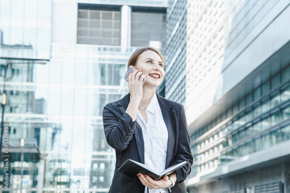 A young woman in a business suit crosses the street. Business woman talking through a mobile phone. A girl with a notepad and a phone near the office. A young woman in a business suit crosses the street. Business woman talking through a mobile phone. A girl with a notepad and a phone near the office.