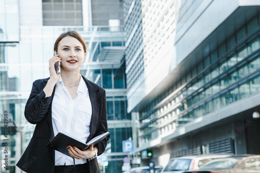 A young woman in a business suit crosses the street. Business woman talking through a mobile phone. A girl with a notepad and a phone near the office. A young woman in a business suit crosses the street. Business woman talking through a mobile phone. A girl with a notepad and a phone near the office.