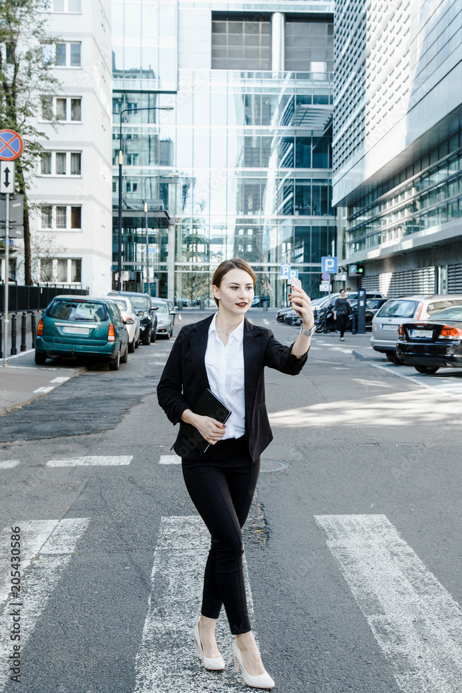 A young woman in a business suit crosses the street. Business woman talking through a mobile phone. A girl with a notepad and a phone near the office.