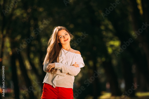 A girl with long hair in stylish clothes embraces herself and looks at the camera lens on the background of the park.