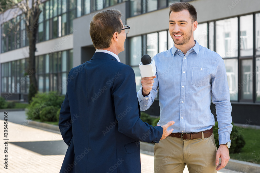 professional smiling journalist interviewing successful mature businessman with microphone