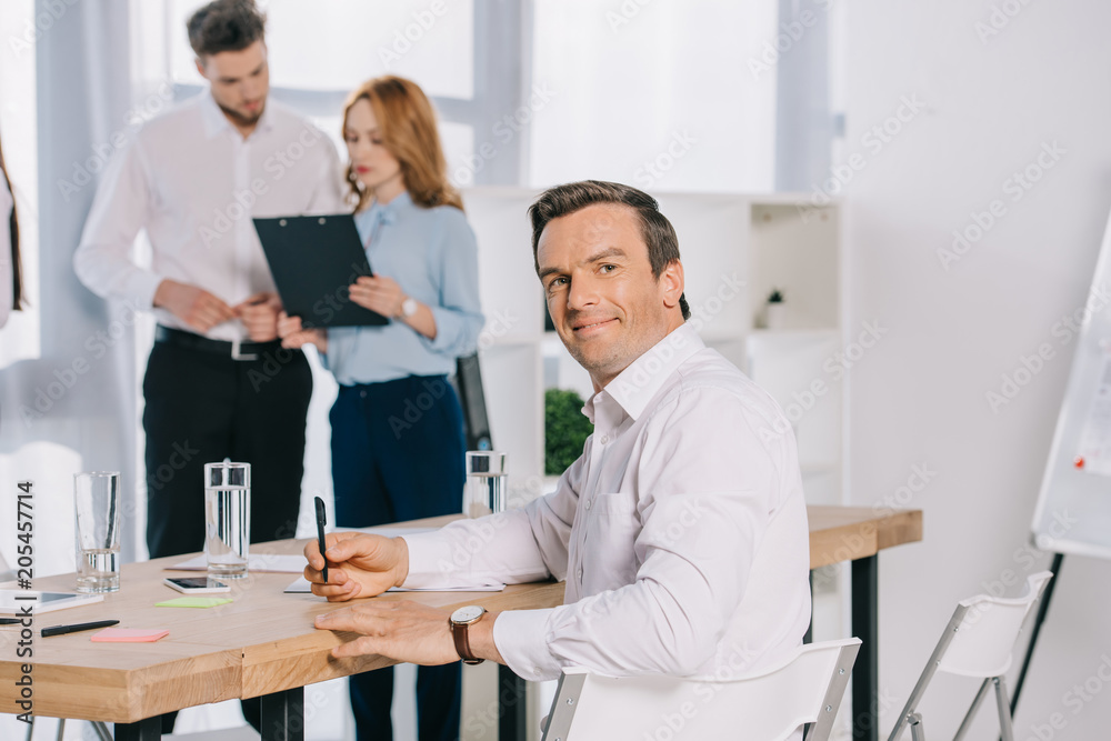 Obraz premium selective focus of smiling businessman at workplace and colleagues discussing work behind in office
