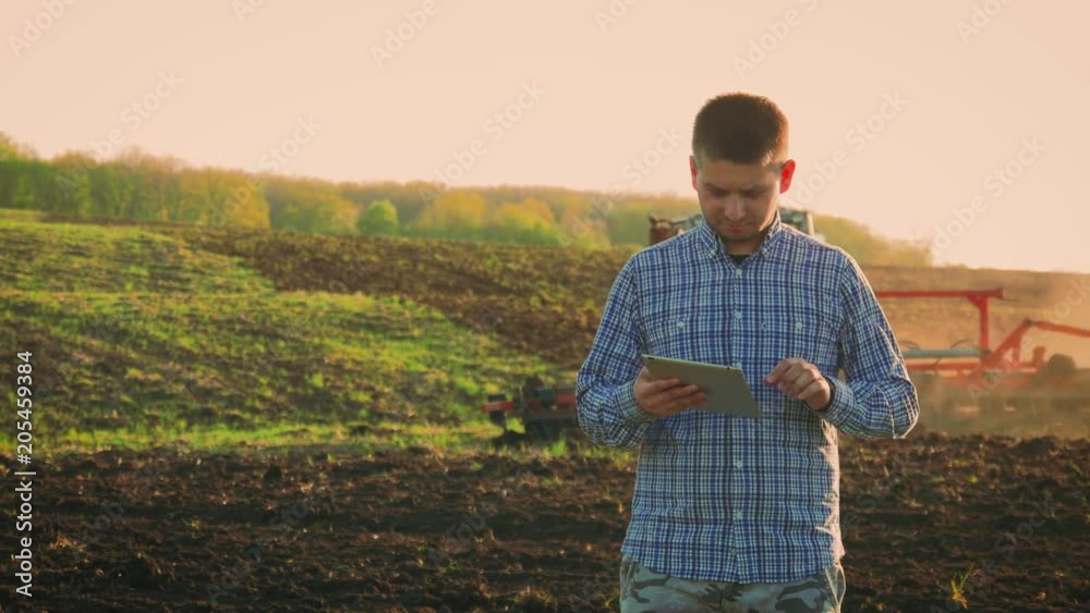 Young farmer holding tablet in front of tractor in field. Farmer controls the work in the field over it's tablet. Seasonal agricultural workers. Young farmer holding tablet in front of tractor in field. Farmer controls the work in the field over it's tablet. Seasonal agricultural workers.