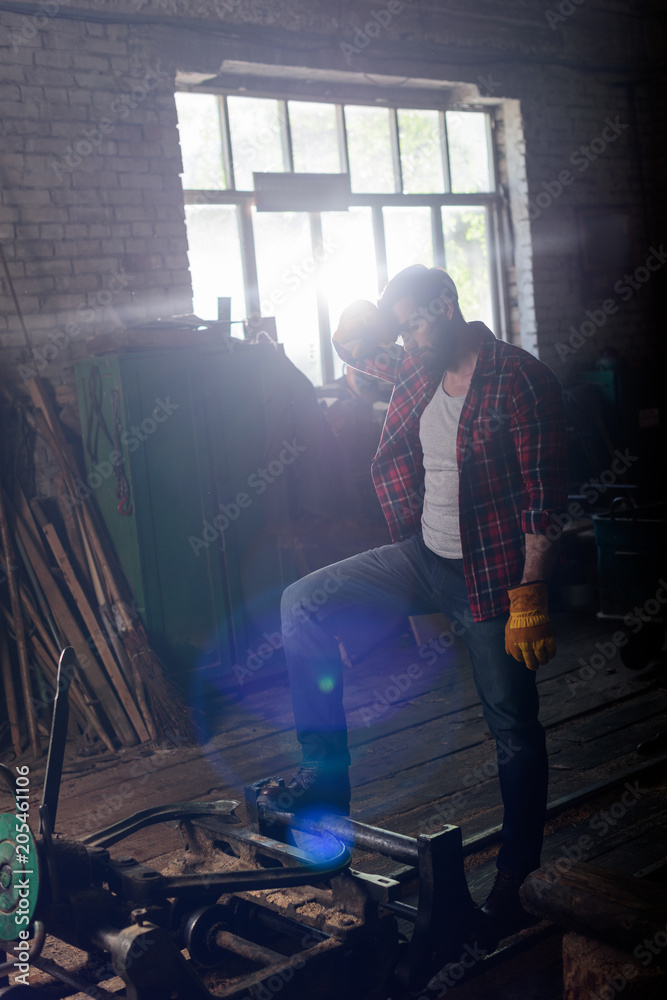 Fototapeta premium bearded worker in protective gloves wiping forehead and standing near machine at sawmill