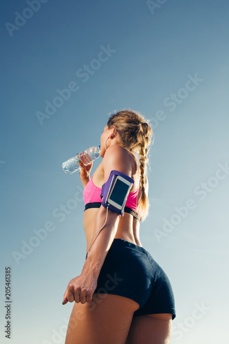 low angle view of sportswoman in earphones with smartphone in running armband case drinking water against blue sky