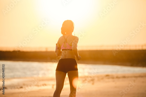 rear view of silhouette of sportswoman in earphones with smartphone in running armband case jogging on beach against sunlight