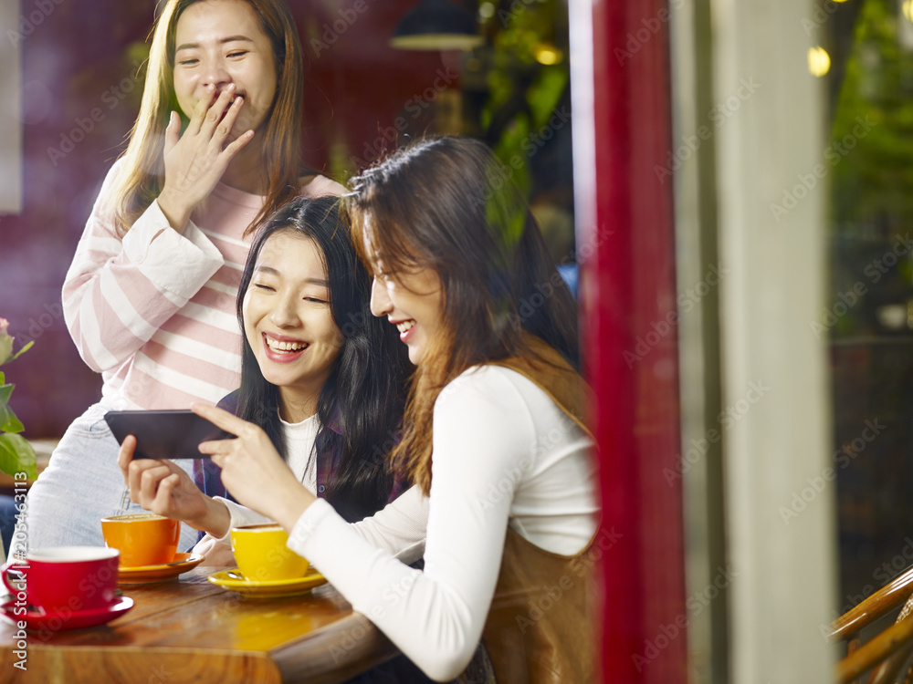 three young asian women looking at mobile phone and laughing in coffee shop three young asian women looking at mobile phone and laughing in coffee shop