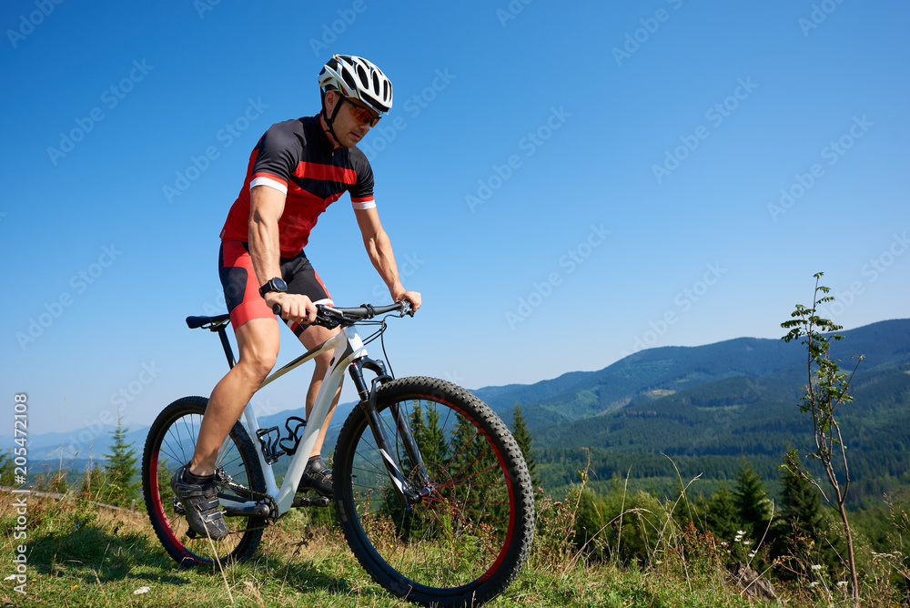 Young professional cyclist in helmet, sunglasses and full equipment riding bike on grassy hill. Mountains and blue summer sky on background. Active lifestyle and extreme sport concept