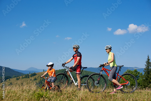 Happy family tourist cyclists, mother, father and child resting with bicycles on the top of grassy hill, looking into distance, on summer sunny day. Active lifestyle, traveling and relations concept