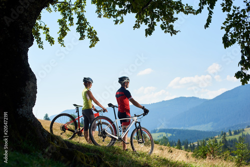 Fototapeta Naklejka Na Ścianę i Meble -  Active couple sportsman cyclists, man and woman in helmets and full equipment, standing with bikes on grassy hill under big green tree branch, enjoying view of blue mountain and sky on background