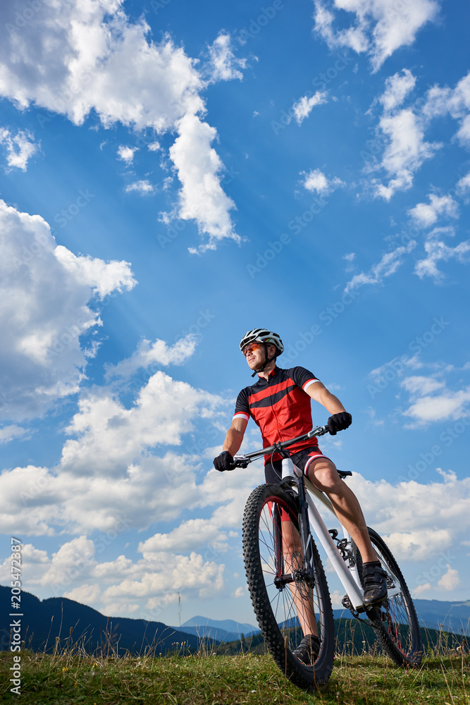 Obraz premium Vertical shot of athletic professional sportsman bicyclist in sportswear and helmet standing with cross country bike, enjoying view of mountains and blue sky with white clouds on sunny summer day