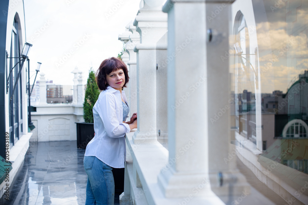 Woman on the balcony in her apartment. Middle age woman enjoying the view standing on the balcony. Rest in city life concept. Woman on the balcony in her apartment. Middle age woman enjoying the view standing on the balcony. Rest in city life concept.