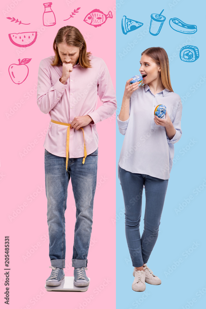 Overweight. Emotional man being on a diet and standing on the scales while his positive wife eating sweets Overweight. Emotional man being on a diet and standing on the scales while his positive wife eating sweets