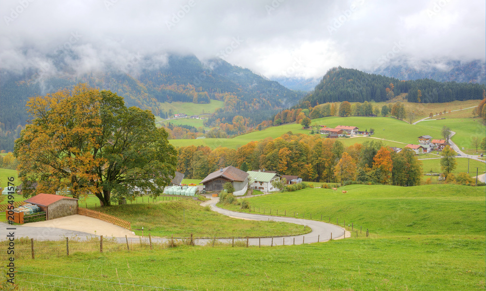 A winding country road curves between green fields and autumn trees leading to a farmhouse on a beautiful hillside ~ Scenery of romantic fall countryside in Ramsau, Bavaria Germany A winding country road curves between green fields and autumn trees leading to a farmhouse on a beautiful hillside ~ Scenery of romantic fall countryside in Ramsau, Bavaria Germany