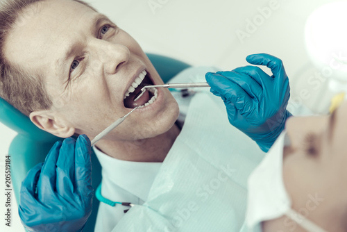 Good teeth. Concentrated responsible dentist wearing blue gloves and examining the teeth of his patient with a help of a little useful dental mirror