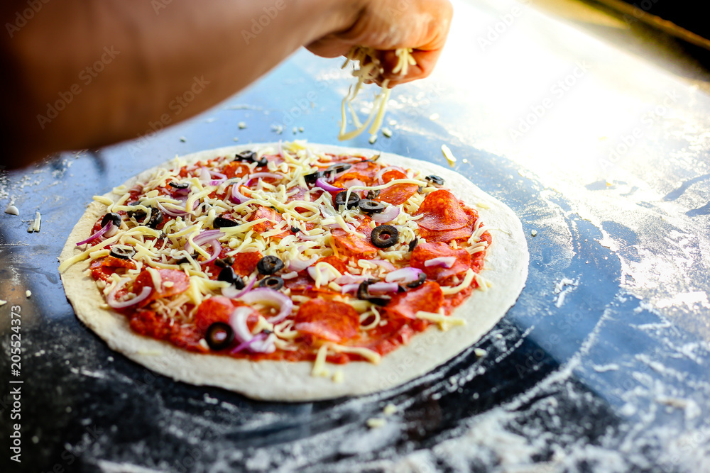 Chef of pizza is garnishing mozzarella cheese on a top or surface of pizza. He is cooking and preparing for baked. The ingredients of pizza have pepperoni, mozzarella cheese, shallots, black olives