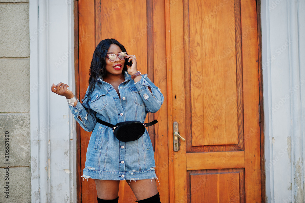 Fototapeta premium African american girl in jeans dress and sunglasses speaking on mobile phone and posed on streets of city. Black stylish model shoot.