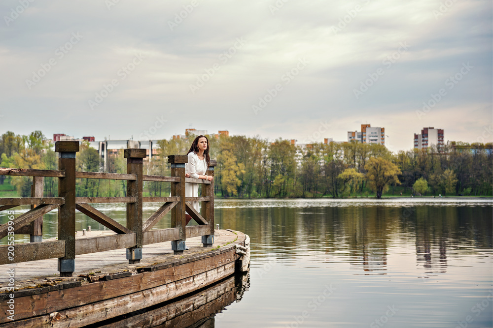 thinking woman in white dress is standing on a wooden pontoon bridge on a river lake with a cityscape on a background 