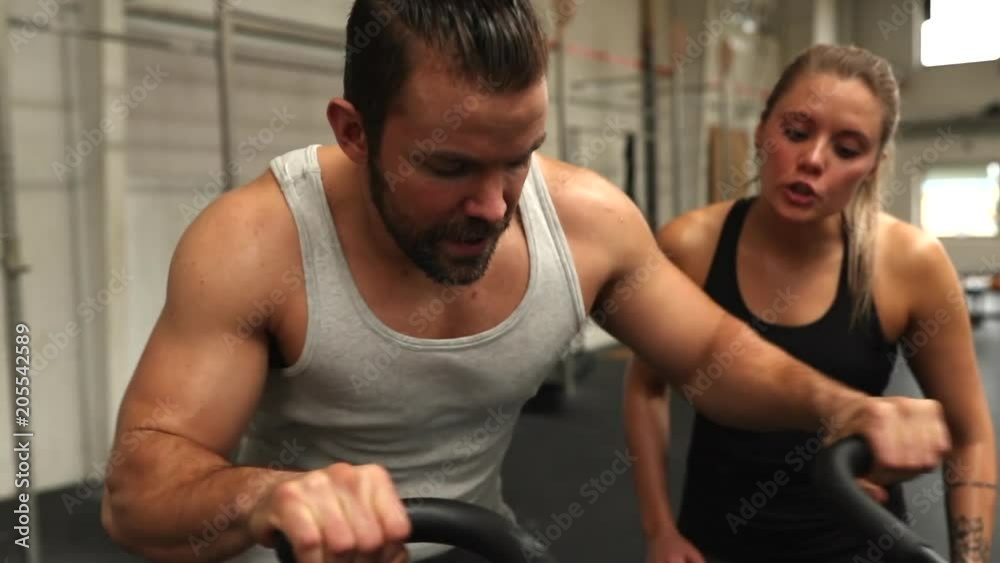 Young man shouting support to his female friend riding a stationary bike during a workout session together at the gym Young man shouting support to his female friend riding a stationary bike during a workout session together at the gym