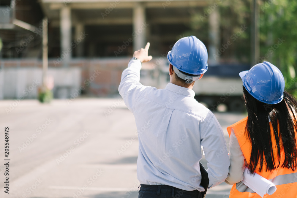 Asian man and woman engineer with the blue safety helmet meeting at the construction site with blueprints and laptop.