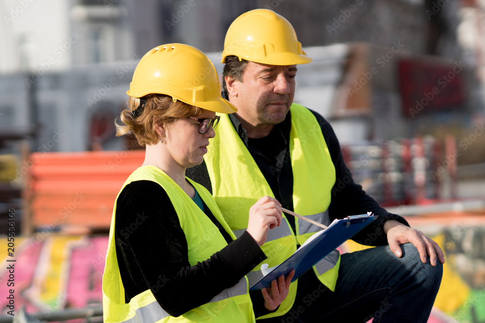 Civil engineers, a man and a woman, wearing safety jackets and helmets, checking projects on construction site Civil engineers, a man and a woman, wearing safety jackets and helmets, checking projects on construction site