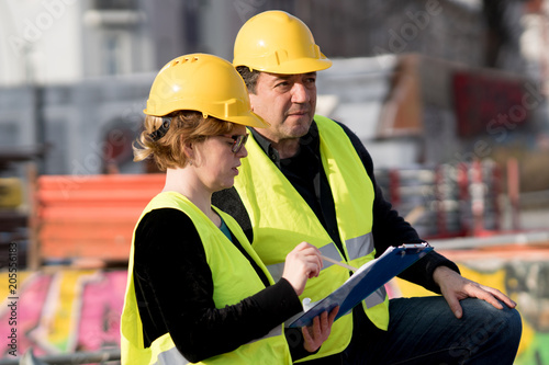 Civil engineers, a man and a woman, wearing safety jackets and helmets, checking projects on construction site