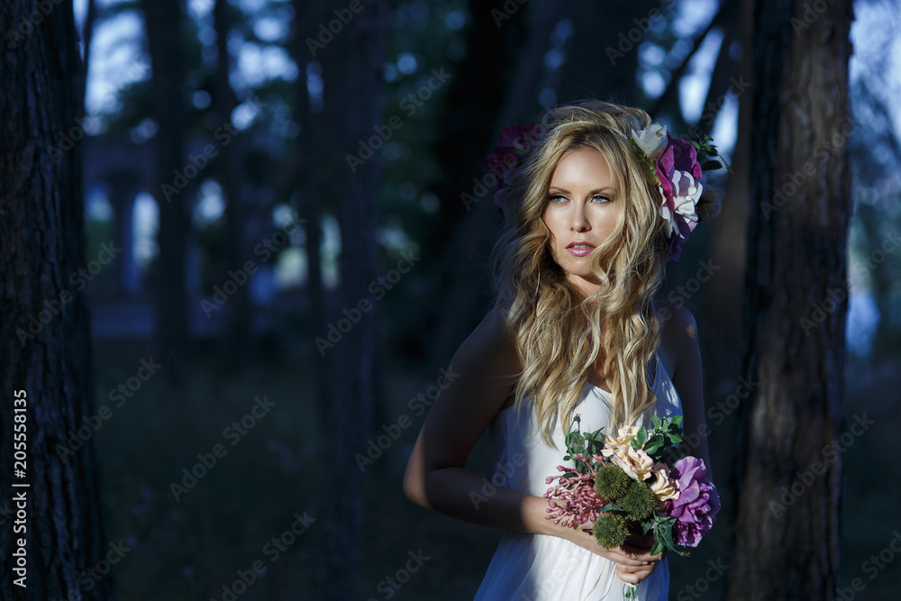 Upper body portrait of young beautiful Caucasian female wearing white dress and flower crown in the dark forest. Posing in romantic style with blue eyes looking at the camera holding a flower bouquet. Upper body portrait of young beautiful Caucasian female wearing white dress and flower crown in the dark forest. Posing in romantic style with blue eyes looking at the camera holding a flower bouquet.