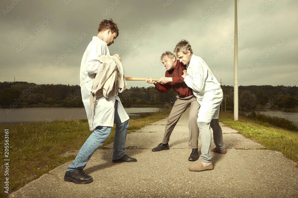 Two older retro paramedic freak hunters in medical coats fighting with crazy man in park Two older retro paramedic freak hunters in medical coats fighting with crazy man in park