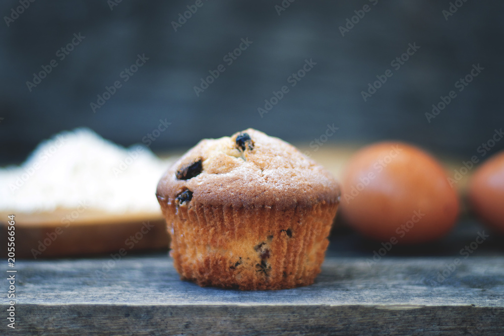 Muffin with raisins sprinkled with powdered sugar on a wooden background and are ingridient on the back of a blurry background Muffin with raisins sprinkled with powdered sugar on a wooden background and are ingridient on the back of a blurry background