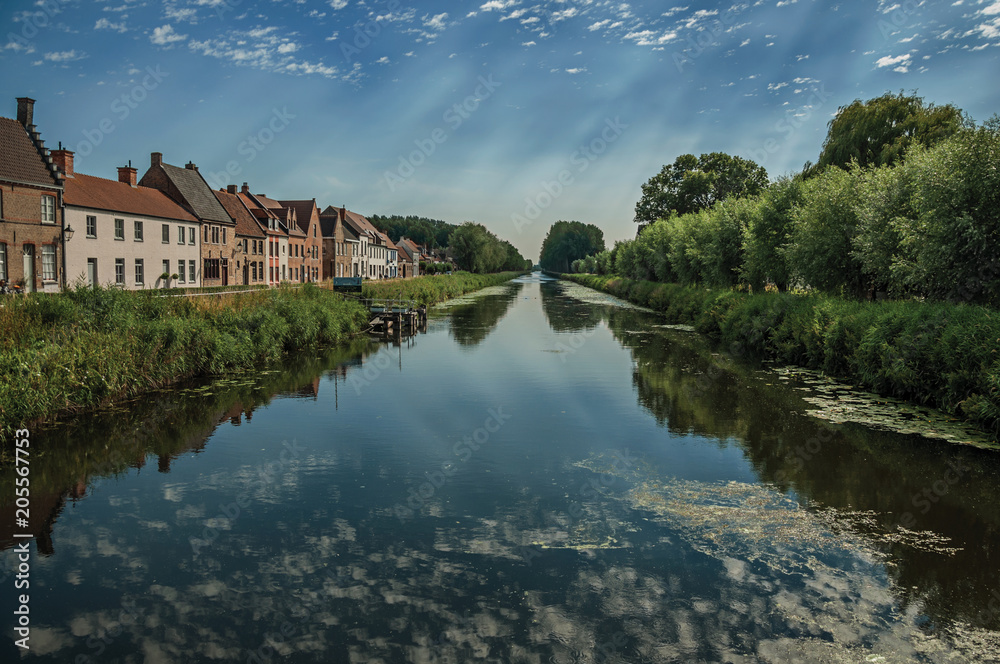 Houses, bushes and grove along canal with sky reflected on water, in the late afternoon and blue sky, near Damme. A quiet and charming countryside old village near Bruges. Northwestern Belgium.
