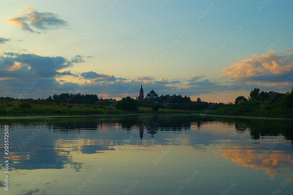 Church in the distance in the forest near the river against the green banks and blue sky with clouds in the evening in the summer reflected in the water