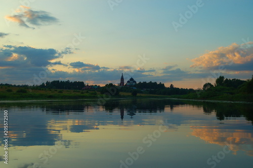 Church in the distance in the forest near the river against the green banks and blue sky with clouds in the evening in the summer reflected in the water