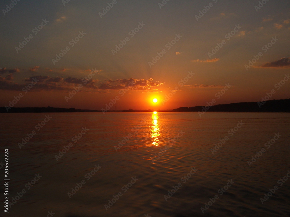A picturesque summer sunset over the water of the lake, bright orange amber sun on the horizon against the background of calm water and evening sky with a small cloud