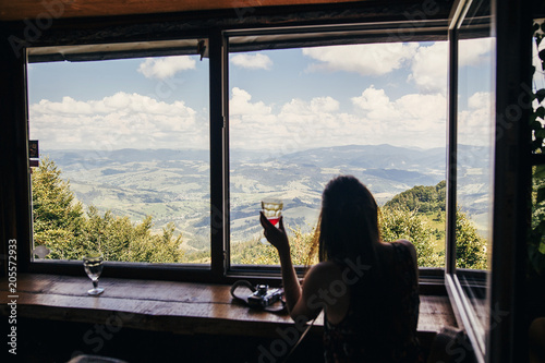stylish girl traveler sitting at window with view on mountains and sky. happy hipster woman holding drink, looking at sky and relaxing. summer vacation concept. focus on mountains