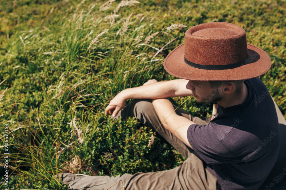 stylish traveler man in hat sitting on top of sunny mountains. travel and wanderlust concept. space for text. happy hipster traveling and camping. summer vacation