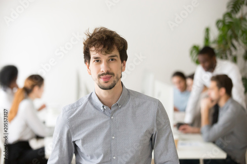 Skilled young professional looking at camera posing in office with team colleagues at background, successful startup founder, corporate employee or sales manager, business leader head shot portrait