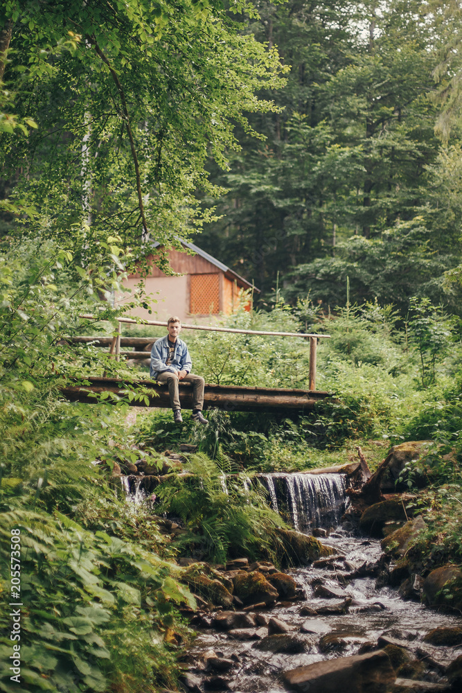 stylish traveler man sitting on wooden bridge at river in sunny forest and mountains. travel and wanderlust concept. space for text. happy hipster guy relaxing in woods. summer vacation